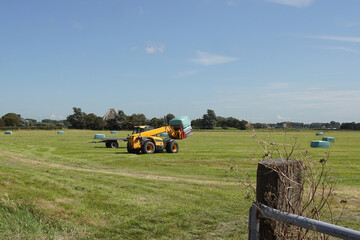 Dutch telescopic handler with bale clamp and behind it a flat trailer to transport the bales. Wrapped hay bales in the meadow. Summer, August, Netherlands.  © Thijs de Graaf