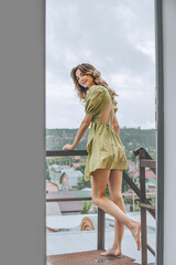 Young model girl in a dress is standing on the balcony against the background of a city and the sky