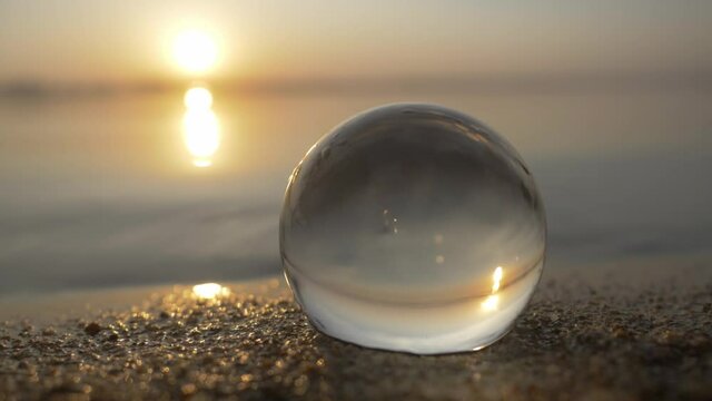 crystal glass ball sphere on sand beach, beautiful sunset on the background. clean planet earth concept. glass transparent ball on blue water background