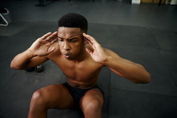 Fototapeta na wymiar Young African American male doing sit-ups on the floor in the gym. Mixed race male personal trainer exercising shirtless indoors. High quality photo