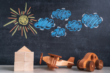 A wooden house and a toy airplane and a car stand against a chalk board with a picture of the sun...