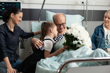 Little granddaughter hugging elderly grandfather visiting him in hospital ward while recovering...