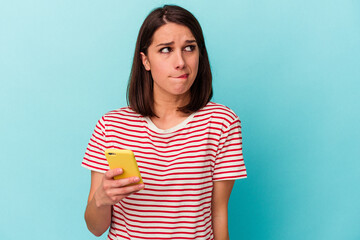 Young caucasian woman holding mobile phone isolated on blue background confused, feels doubtful and unsure.