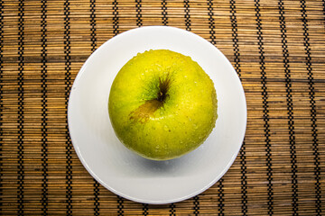 big ripe yellow green apple with brown line and water drops on a white small plate bamboo mat