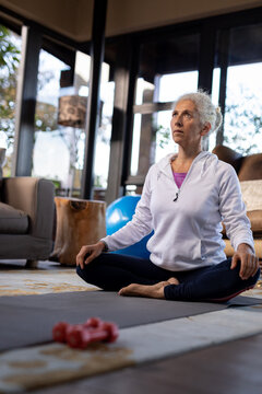 Senior caucasian woman in living room exercising, sitting on the floor and meditating