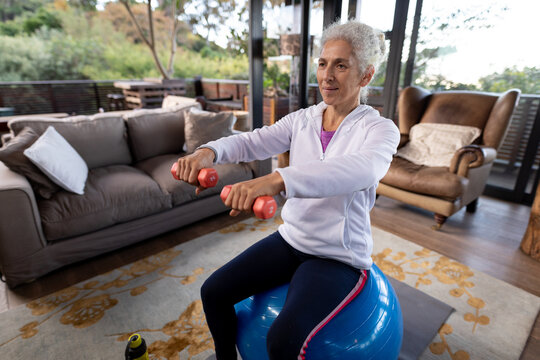 Happy senior caucasian woman in living room exercising, sitting on swiss ball lifting dumbbells