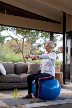 Senior caucasian woman in living room exercising, sitting on swiss ball lifting dumbbells