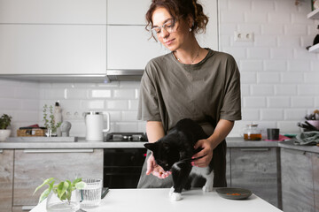 Beautiful young woman wearing casual home t shirt feeding cute cat on the kitchen
