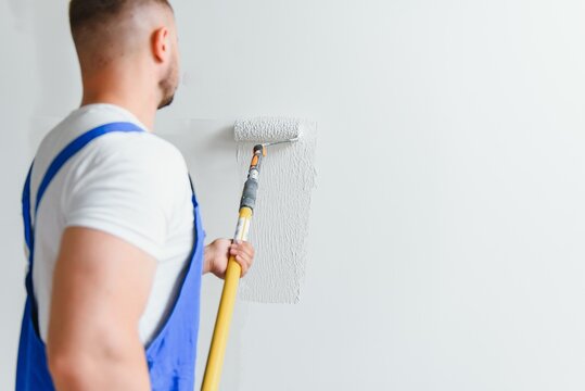 Workman in blue overalls painting the wall with a roller, view from the side