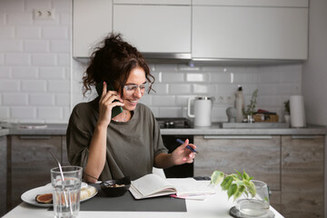 business woman wearing casual home t shirt doing paper works on the kitchen while having breakfast, making notes to daybook