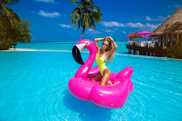 Beautiful sexy tanned woman bikini model on pink flamingo in pool. Young glamour girl in swimsuit...