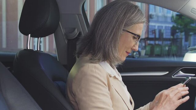 Mature woman sitting in car and chatting with friends on smartphone, dating app