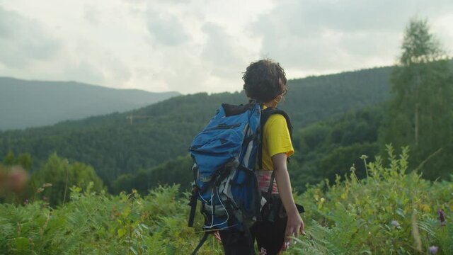 Portrait of smiling charming african american female traveler with backpack walking through mountain meadow, touching wild plants and flowers, outstretching hand asking to follow her during hiking.