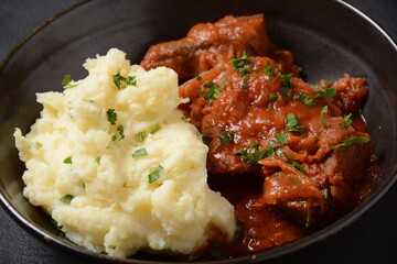 Stewed beef pieces in sauce with mashed potatoes and herbs on dark background