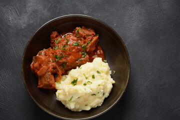 Stewed beef pieces in sauce with mashed potatoes and herbs on dark background