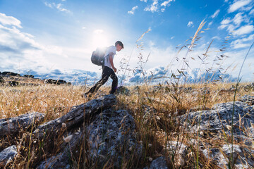 Side view of a teenager carrying backpack hiking through rocky valleys with the sun behind him and...