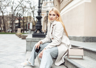 Young fashion blonde woman in a trench coat sits on stairs in the city and holding smartphone
