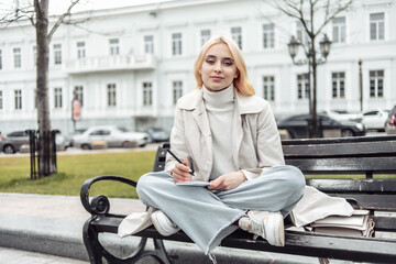 Stylish young woman tsudent sits on a bench and writes in notebook