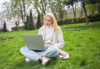 Young blonde woman uses a laptop while sitting on the grass in the park