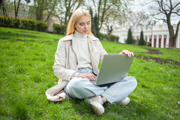Young blonde woman uses a laptop while sitting on the grass in the park