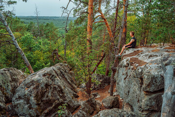 A young man is sitting on the edge of a cliff overlooking a grand panorama of a pristine forest with rocks