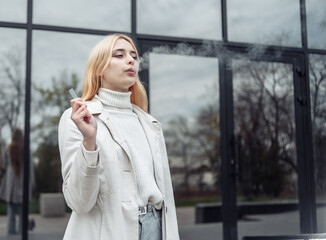 Young stylish blonde woman smokes electronic cigarette near the business center