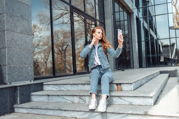 Young pretty woman in headphones and with a smartphone sits on the stairs near the business center