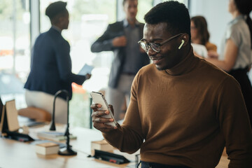 Obraz premium Cheerful African-American Business Man Enjoying Listening to Music on Wireless Earphones and Phone while Leaning on the Table in a Open Plan Office with Multi-Ethnic Team Talking in the Background