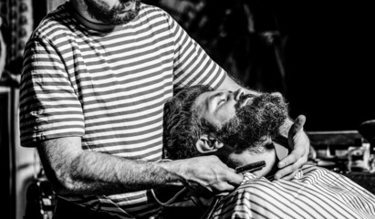 Bearded male sitting in an armchair in a barber shop while hairdresser shaves his beard with a...