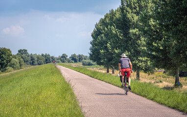 Fototapeta premium Man in shorts and hat cycles alone on a Dutch dike. It is a sunny day in the summer season. The photo was taken in the province of North Brabant.