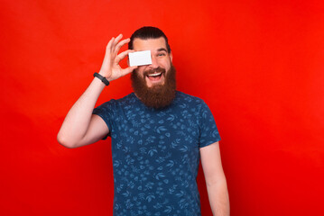 Happy bearded man is covering his eye with a credit or contact card over red background.