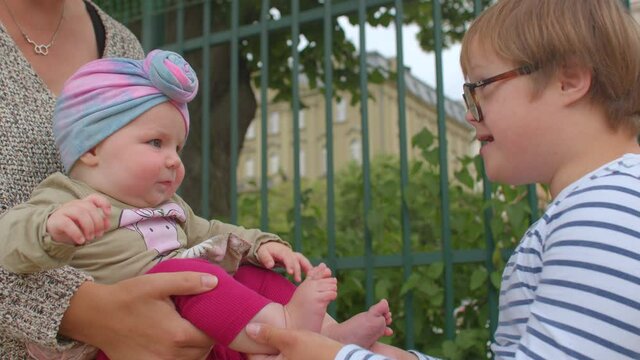 Down syndrome boy touches and examines baby toddler feet. Acquaintance with the new, knowledge of the world. The girl in her mother's arms rejoices at the attention.