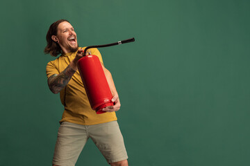 Comic portrait of young man with fire extinguisher having fun isolated on green studio background....