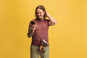 Fototapeta premium Portrait of young man, photographer, cameraman with retro camera using phone isolated on yellow studio background. Concept of occupation, job, funny meme emotions