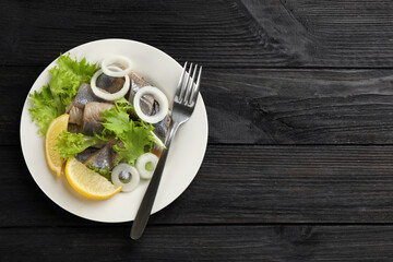 Sliced salted herring fillet served with lettuce, onion rings and lemon on dark wooden table, top view. Space for text
