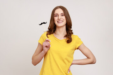 Portrait of funny playful wavy haired teen girl in urban style T-shirt keeping fake paper mustache and looking at camera with happy expression. Indoor studio shot isolated on gray background.