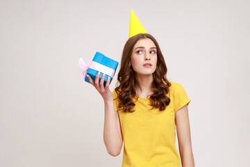 Curious adorable woman of young age in yellow casual T-shirt shaking blue giftbox near ear trying to guess her present, expectating expression. Indoor studio shot isolated on gray background.