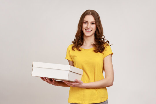 Delivery and cargo transportation. Young attractive female holding cardboard box, looking smiling to camera. Courier carrying parcel. Indoor studio shot isolated on gray background.