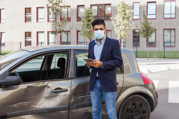 Young adult brunette man in surgical medical mask, jacket and jeans posing on city street with...
