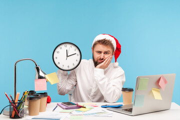 Unhappy man office worker in santa claus hat and white shirt holding wall clock, celebrating new...