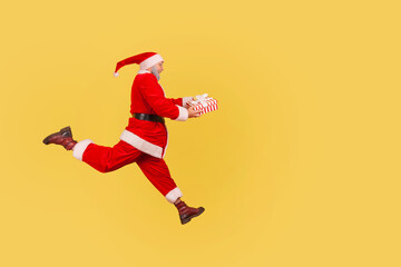 Side view full length of elderly man with beard in santa claus costume running with present box in hands, being hurry to congratulate with New year. Indoor studio shot isolated on yellow background.