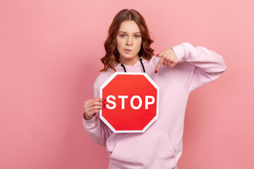 Portrait of displeased curly haired teenage girl in hoodie pointing finger at stop sign, strictly looking camera. Indoor studio shot isolated on pink background