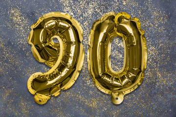 The number of the balloon made of golden foil, the number ninety on a gray background with sequins. Birthday greeting card with inscription 90. Anniversary concept. Celebration event, template.