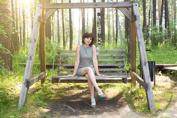 A happy brunette in a striped dress rides on a swing in a birch grove