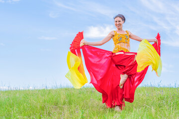 Young woman in a bright national costume is dancing an oriental dance with fans on a green meadow against a blue summer sky. Pleasant pastime.