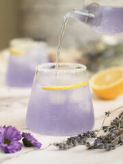 Glass of fresh lavender-lemonade soda drink, tea pot pouring into a glass, salt&sugar on the edge with lemon in background. A fresh healthy organic drink ideal for summer and full of health benefit
