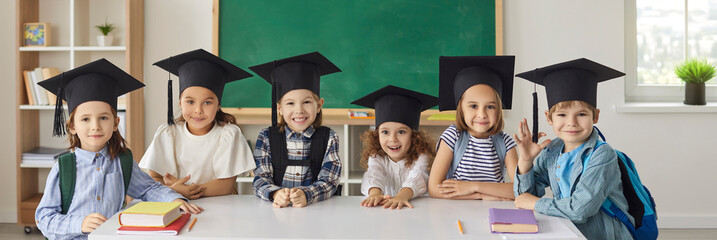 Website banner with group portrait of cute school children in mortarboards. Happy little kids in academic caps smiling and looking at camera standing by table with books in new modern classroom
