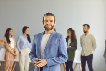 Portrait of a smiling and confident young business man on the background of employees. Stylish bearded man in a blue jacket and white T-shirt looks at the camera while standing in a bright room.