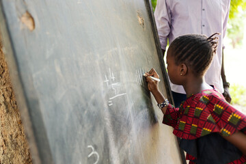 Close up side view of a little African schoolgirl writing tally marks on a blackboard with a big...