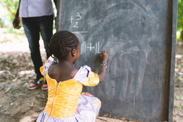 Rear view of a black African firstgrader kneeling in front of a blackboard writing numbers during...